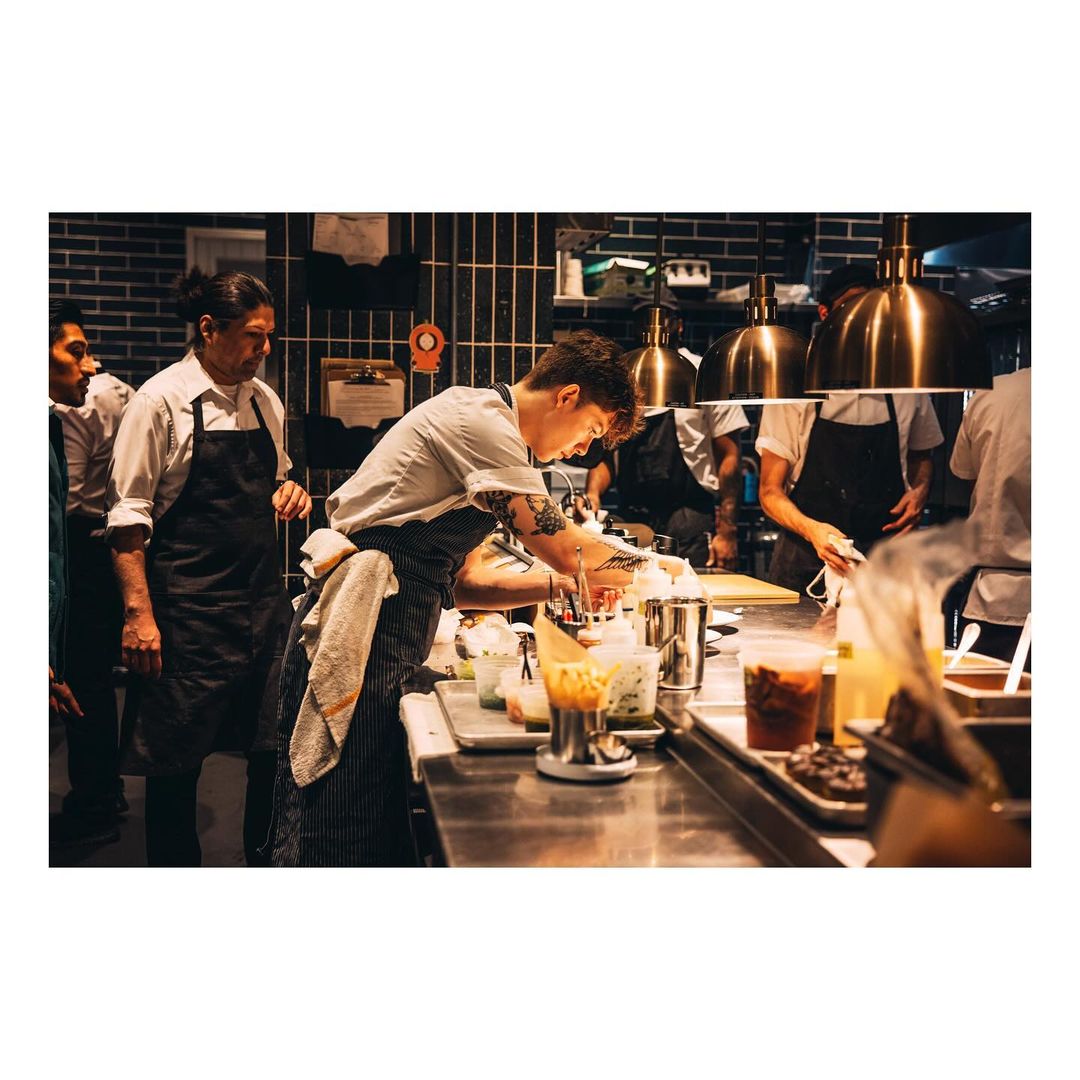 Busy commercial kitchen scene with chefs preparing food at stainless steel counters under bright lighting and exhaust hoods.