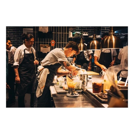 Busy commercial kitchen scene with chefs preparing food at stainless steel counters under bright lighting and exhaust hoods.