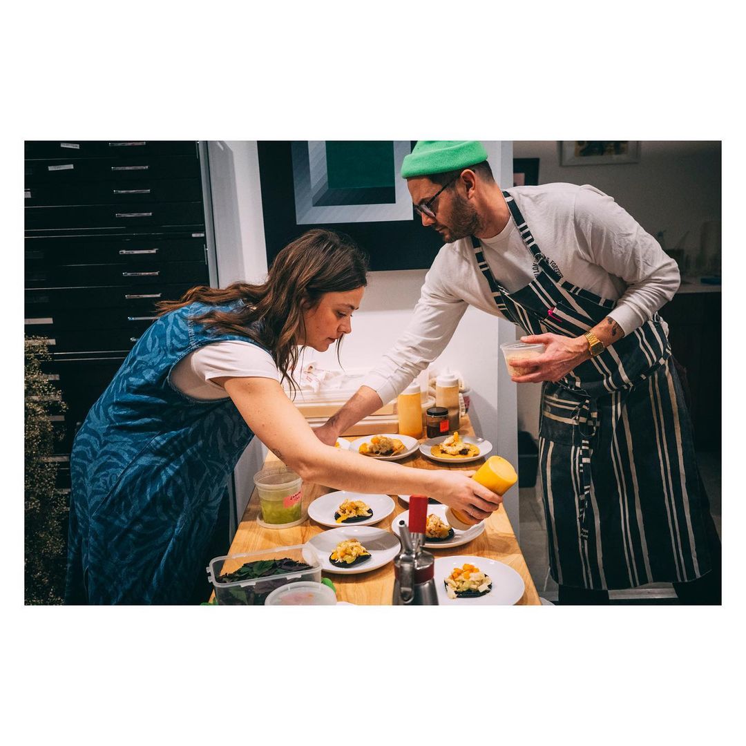 Two people prepare food in a bright indoor setting. A woman in a blue dress and a man in a green beanie plate food with sauces.