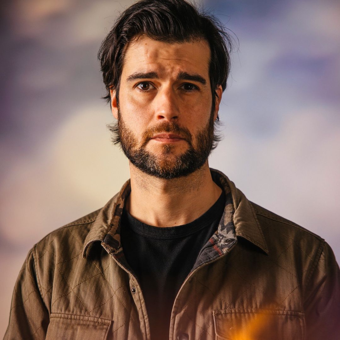 Portrait of an adult male with dark hair and a beard wearing a casual shirt, gazing at the camera with a neutral expression.
