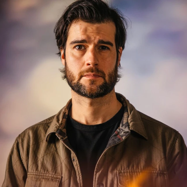 Portrait of an adult male with dark hair and a beard wearing a casual shirt, gazing at the camera with a neutral expression.