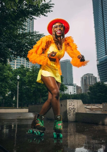 An African-American woman in a colorful outfit is joyfully roller skating outside on a wet surface near city buildings and green trees.