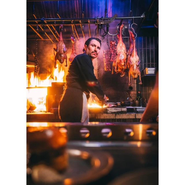 Professional chef cooking food over open fire in commercial kitchen, with hanging game birds and meats; dark and moody lighting.