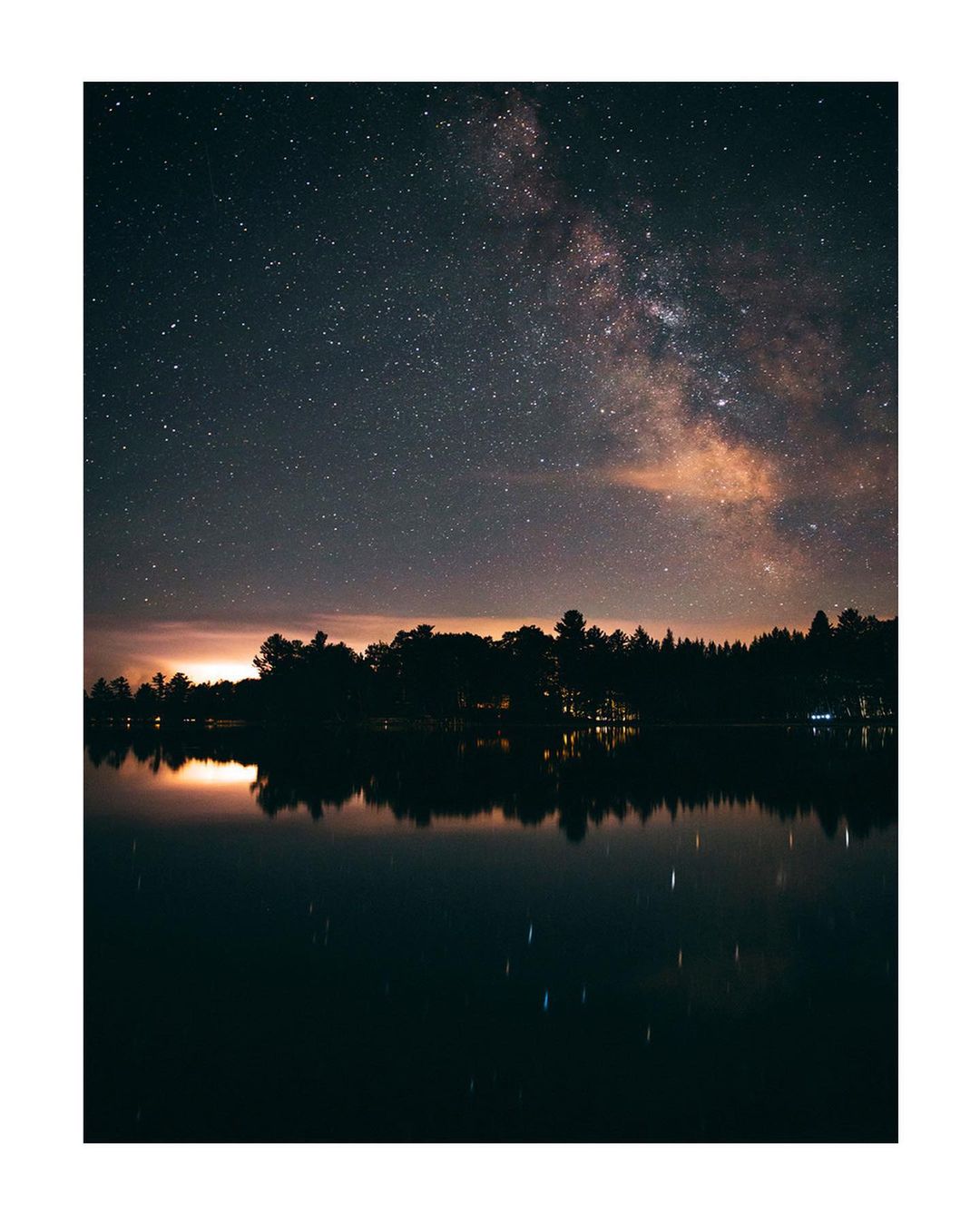 Night sky landscape features the Milky Way and stars above a tranquil body of water and silhouetted trees under the glow of the night.