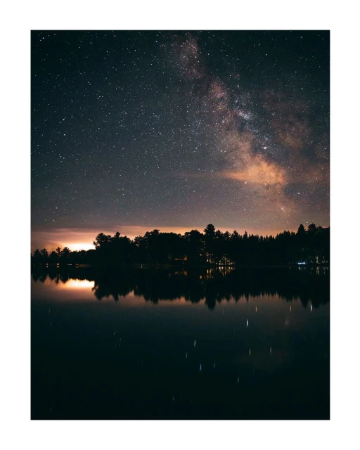 Night sky landscape features the Milky Way and stars above a tranquil body of water and silhouetted trees under the glow of the night.
