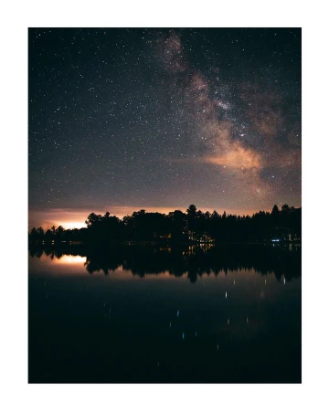 Night sky landscape features the Milky Way and stars above a tranquil body of water and silhouetted trees under the glow of the night.