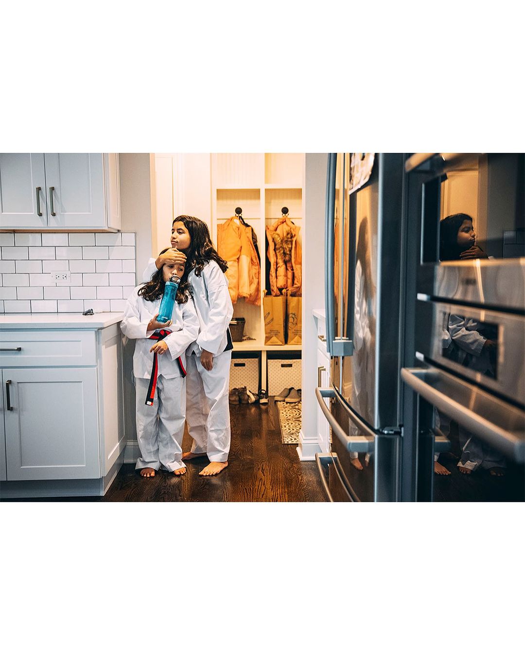 Two young female martial artists in white uniforms stand in a modern kitchen, one holding a blue water bottle, looking off-camera.
