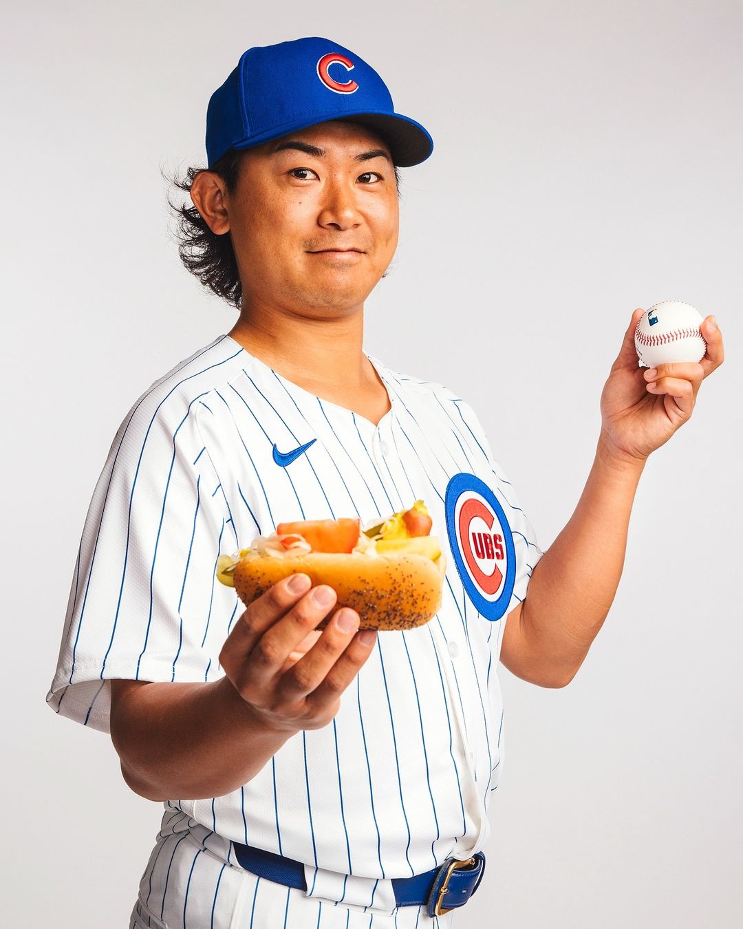 A smiling adult male wearing a Chicago Cubs baseball uniform and cap, holding a hotdog in one hand and a baseball in the other, on white.