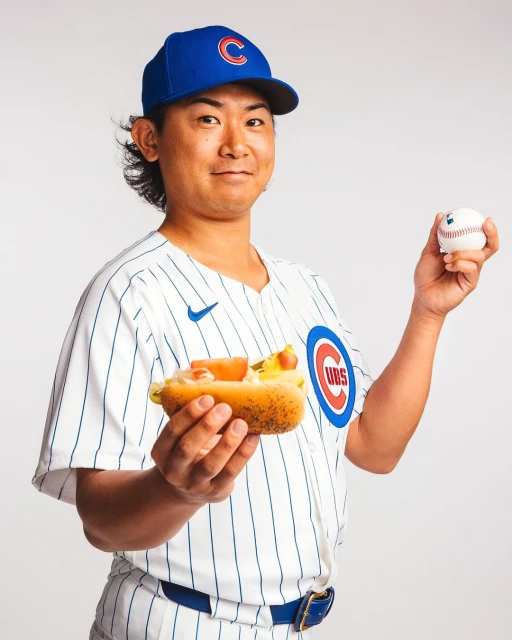 A smiling adult male wearing a Chicago Cubs baseball uniform and cap, holding a hotdog in one hand and a baseball in the other, on white.
