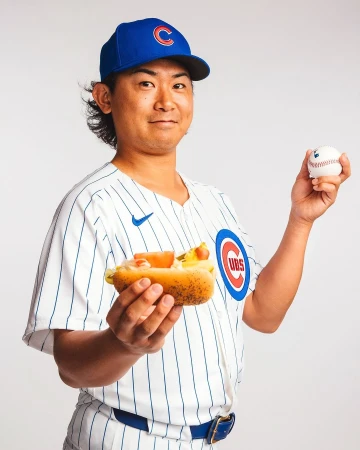 A smiling adult male wearing a Chicago Cubs baseball uniform and cap, holding a hotdog in one hand and a baseball in the other, on white.