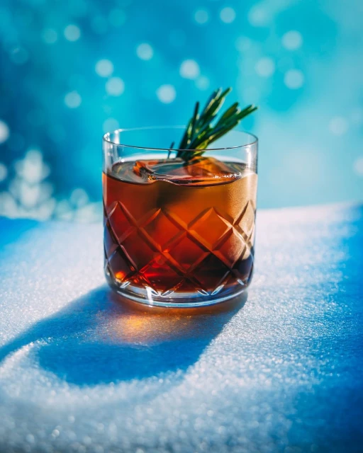Crystal glass of amber liquid with ice and rosemary garnish on a snowy surface, set against a blurred blue background.