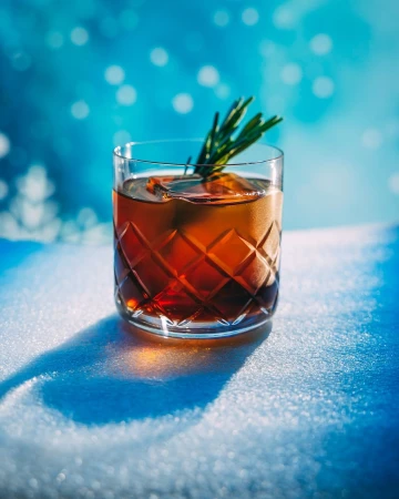 Crystal glass of amber liquid with ice and rosemary garnish on a snowy surface, set against a blurred blue background.