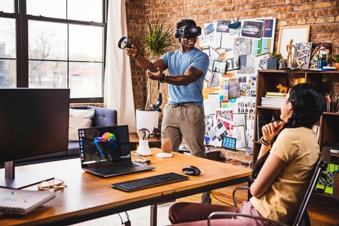 A man plays VR games in an apartment, wearing a VR headset and holding controllers, with a woman watching and office equipment nearby.
