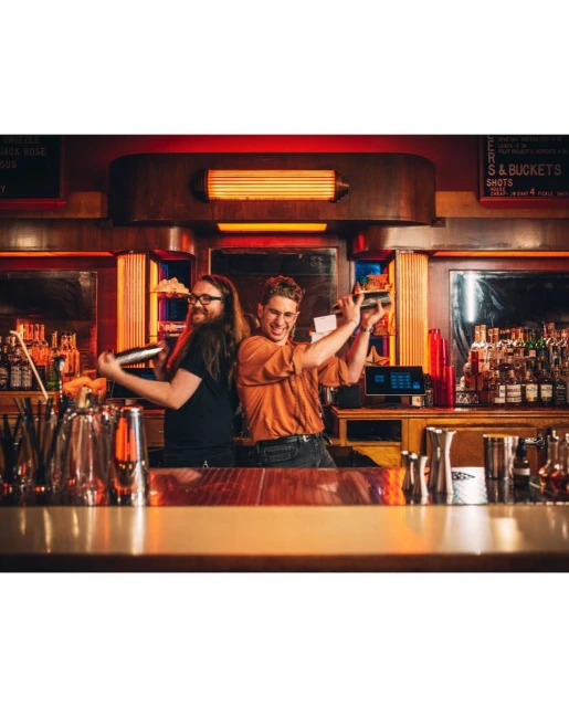 Two bartenders mixing drinks behind a bar with bottles and bar equipment, creating an energetic atmosphere with a warm, red lighting.