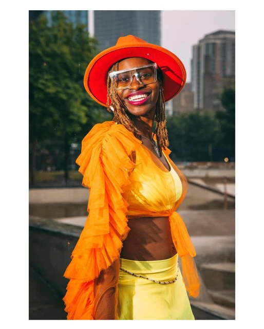 Smiling woman in a city park wearing an orange hat and top, transparent glasses, and yellow skirt posing in front of trees and buildings.