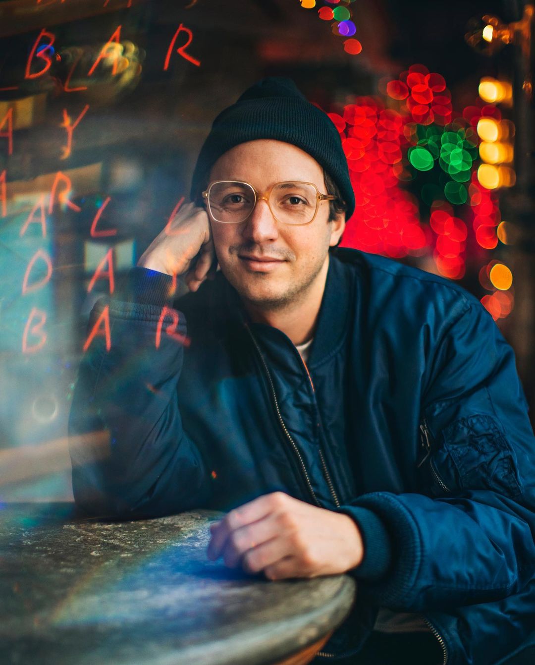 Smiling man with glasses and beanie poses indoors in front of blurred bar window, wearing a dark blue jacket, medium shot.