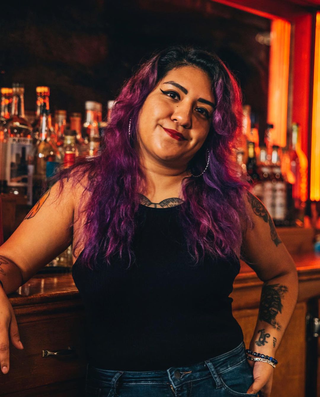 Portrait of a woman with purple hair standing at a bar, featuring liquor bottles and warm lighting in an indoor setting.