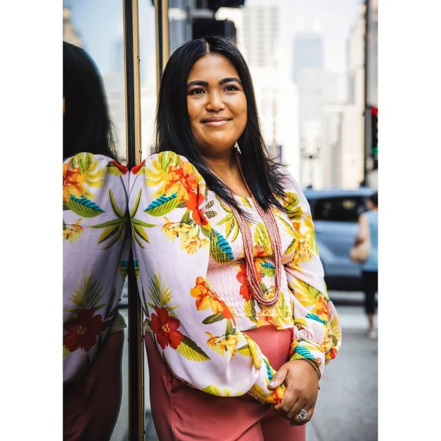 Woman with floral blouse leans against glass building, smiling in a medium shot with skyscrapers and a city street in the background.