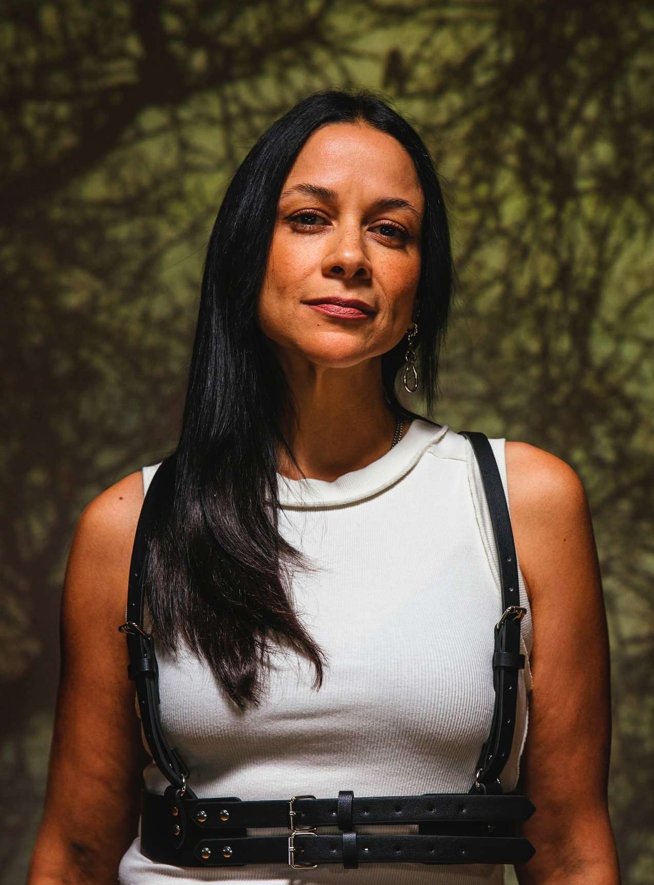 Portrait of a woman with long dark hair wearing a white shirt and black harness against a shadowed, neutral background.