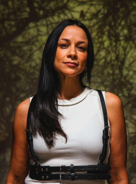 Portrait of a woman with long dark hair wearing a white shirt and black harness against a shadowed, neutral background.