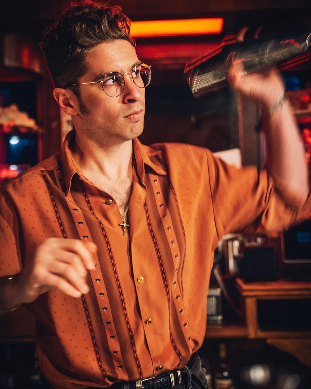 Stylish man in an orange shirt mixes a drink with a cocktail shaker in a dimly lit bar setting with vintage vibes and red lighting.
