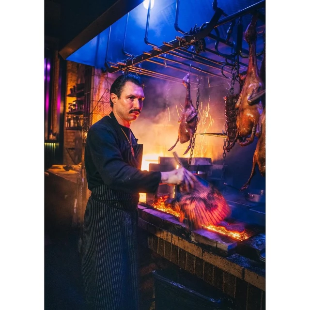 Chef tending meat roasting on a fire grill inside a commercial kitchen, wearing an apron and black top in a low-lit environment.