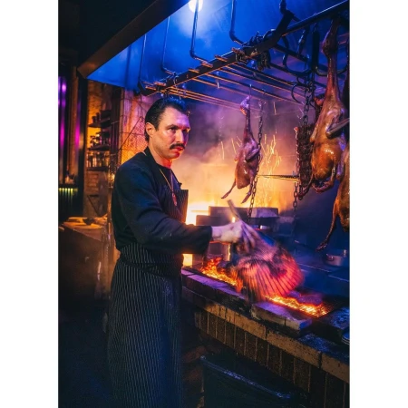 Chef tending meat roasting on a fire grill inside a commercial kitchen, wearing an apron and black top in a low-lit environment.