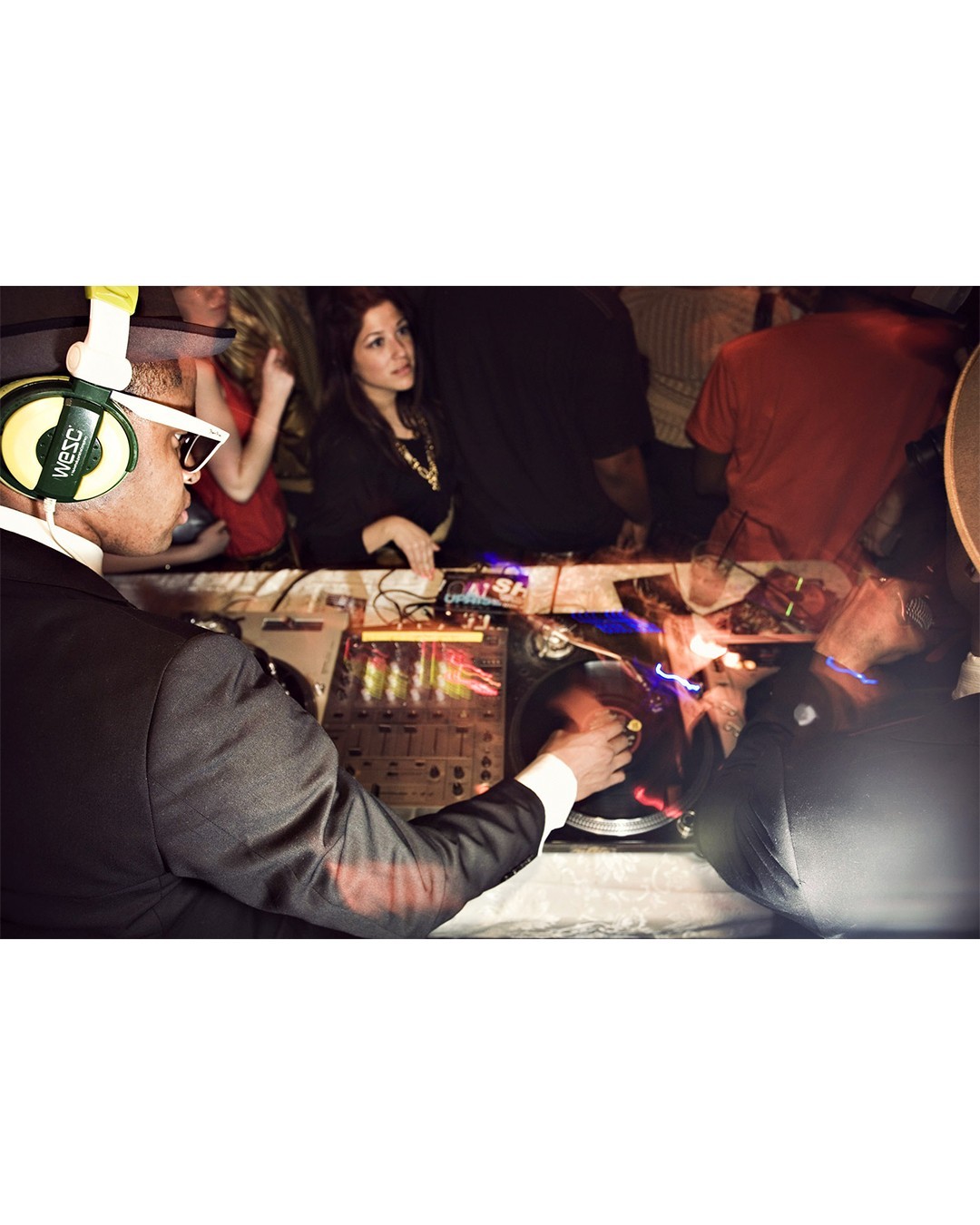 Male DJ in a suit plays music with DJ mixing console, wearing headphones and glasses, at an event with partygoers in the background.