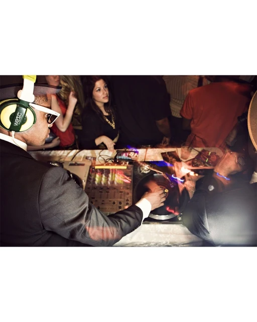 Male DJ in a suit plays music with DJ mixing console, wearing headphones and glasses, at an event with partygoers in the background.