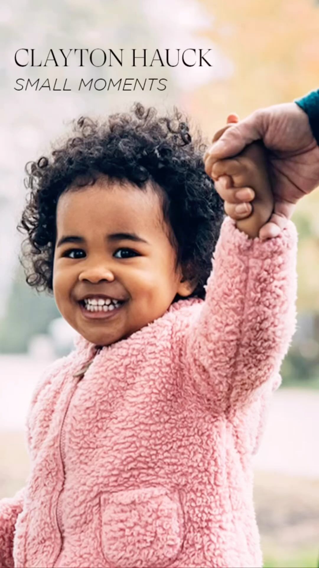 Close-up of a happy toddler with curly hair, wearing a pink sherpa jacket, being held by an adult with a blurred outdoor background.