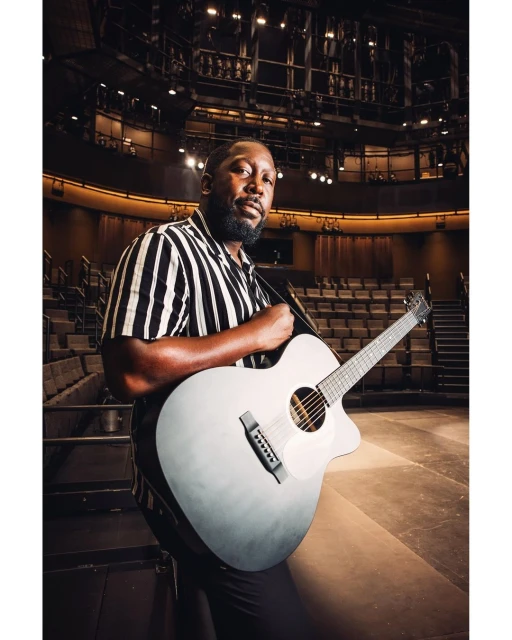 A man with a guitar stands in a theater, wearing a black and white striped shirt and dark pants. The theater seats are visible in the background.