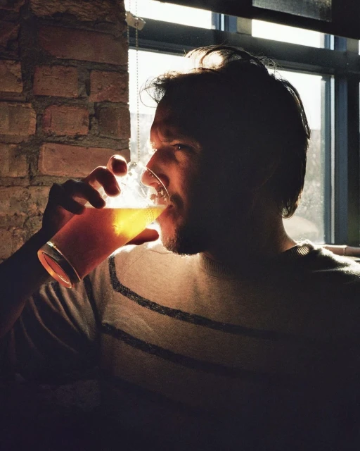 Man drinks beer from a glass in a dimly lit room with a brick wall and window in the background, illuminated by backlighting.