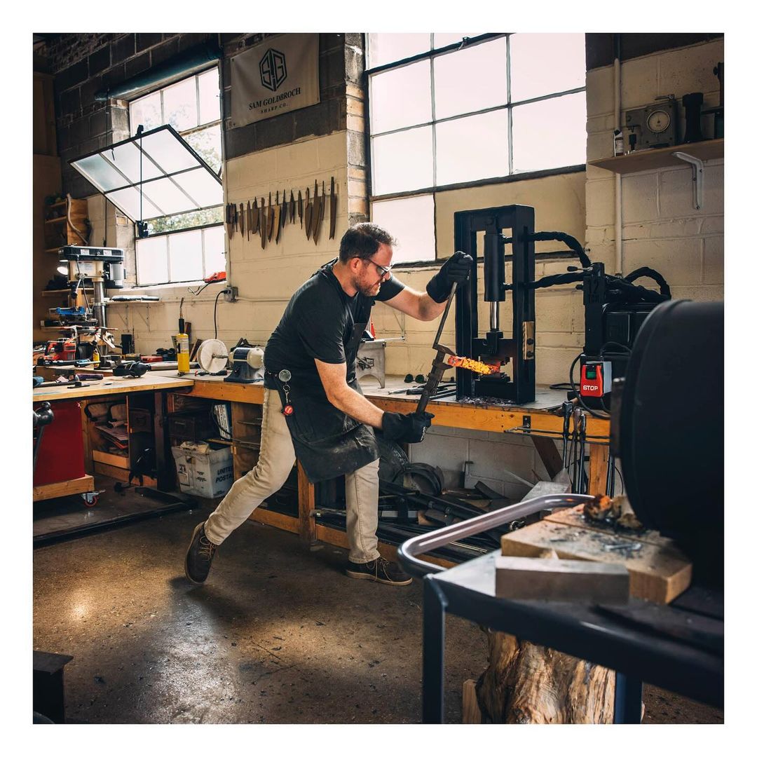 A male blacksmith works in his metal shop. He is using a machine to shape a glowing hot piece of metal. Tools line the wall.