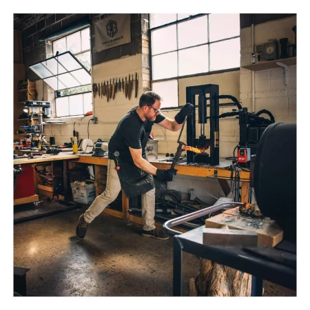 A male blacksmith works in his metal shop. He is using a machine to shape a glowing hot piece of metal. Tools line the wall.