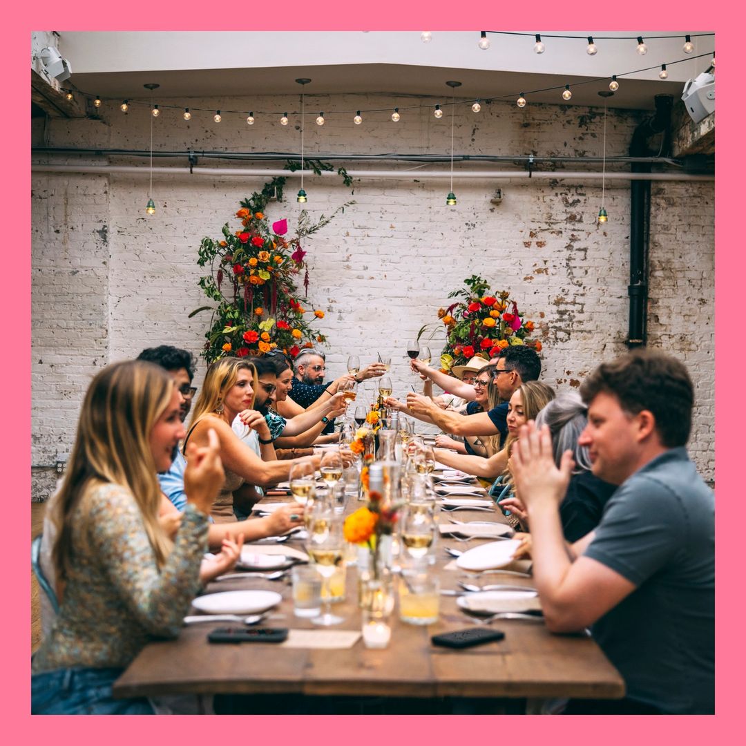 A long table with a group of people raising glasses for a toast indoors under string lights and floral arrangements for a celebratory gathering.