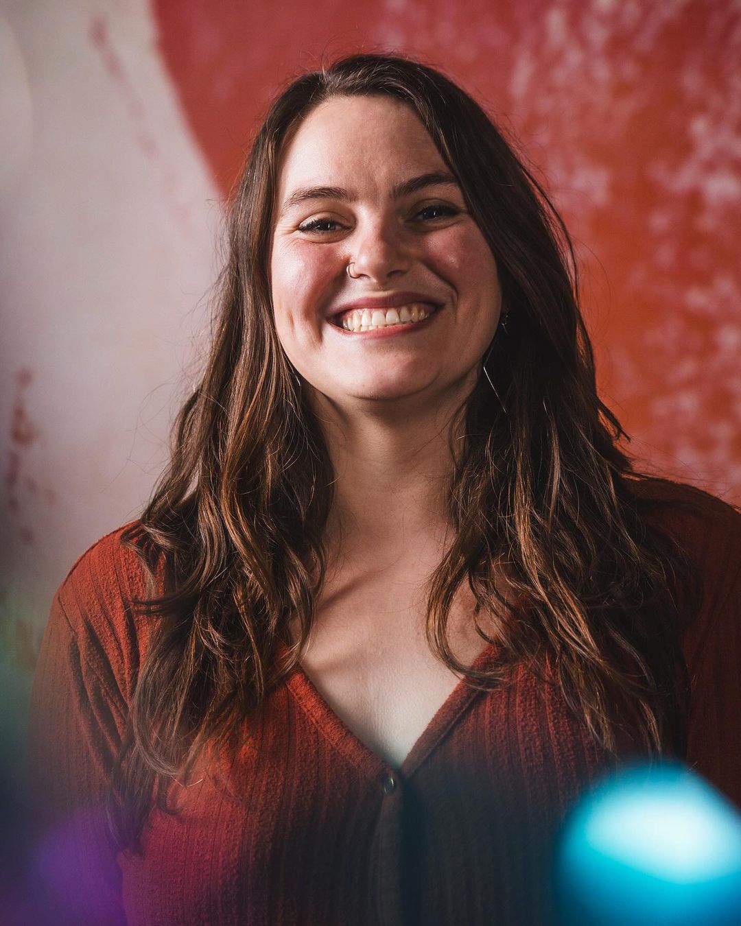 Smiling woman with long brown hair, wearing an orange top, poses indoors with an abstract red and white background, shot in a medium close-up.