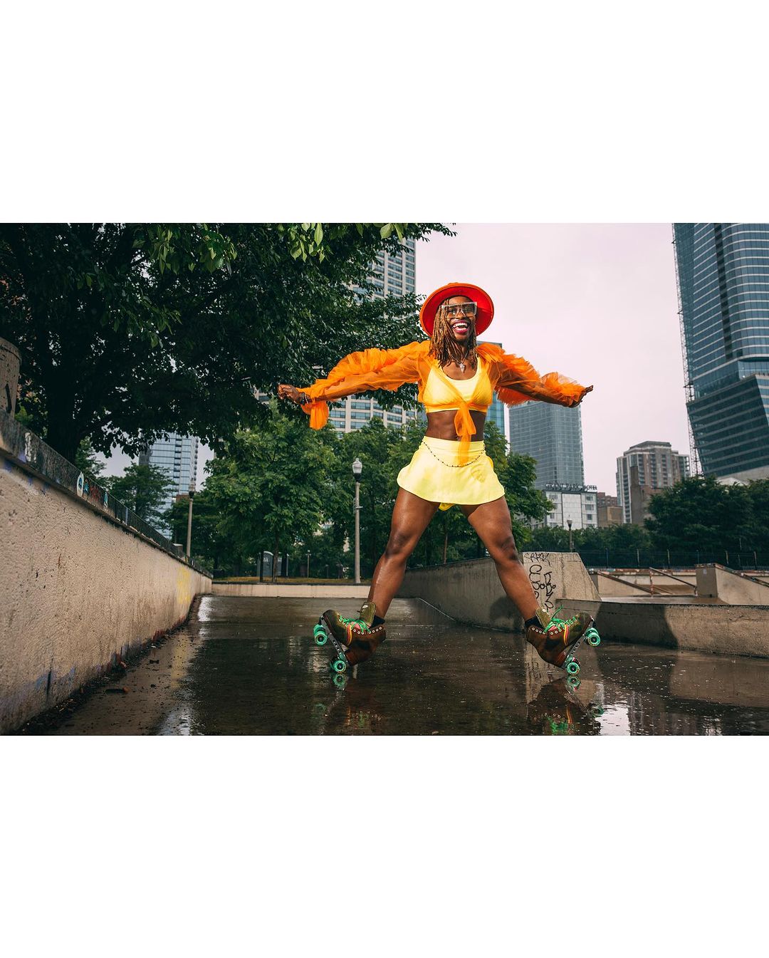 Adult female roller skater in a yellow skirt and crop top poses confidently in an urban skate park with skyscrapers in the background.