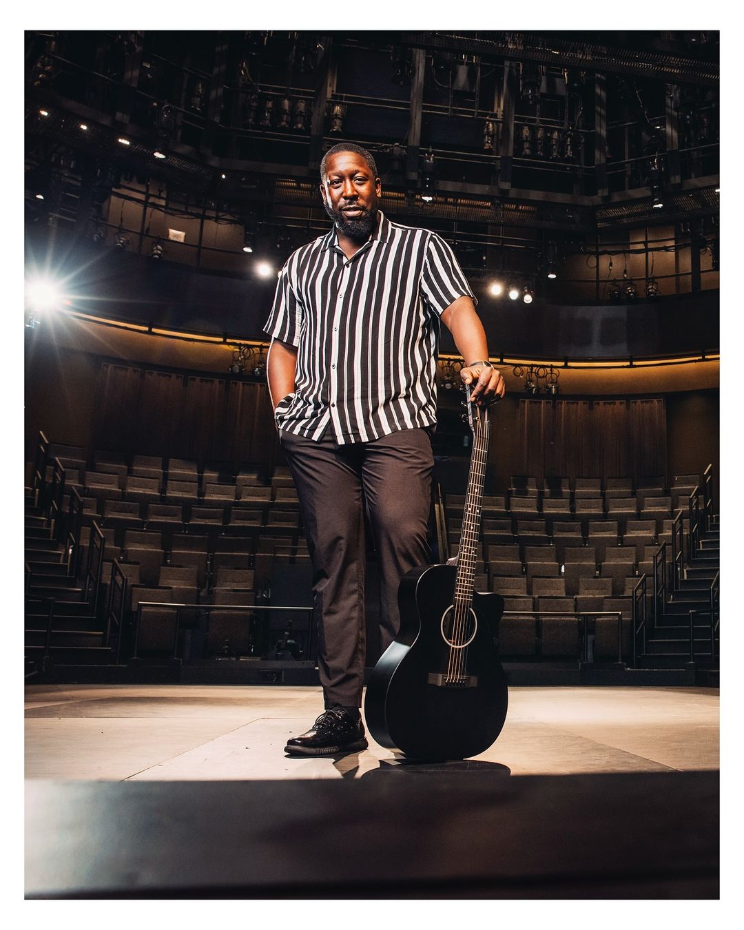 Man stands with guitar on stage in theater. Adult male with short beard is wearing vertically striped shirt and dark pants.