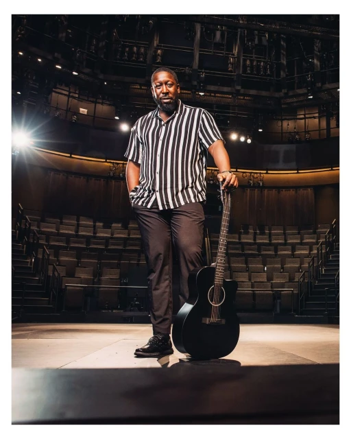 Man stands with guitar on stage in theater. Adult male with short beard is wearing vertically striped shirt and dark pants.