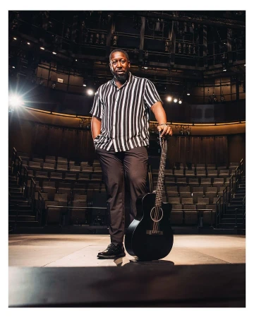 Man stands with guitar on stage in theater. Adult male with short beard is wearing vertically striped shirt and dark pants.