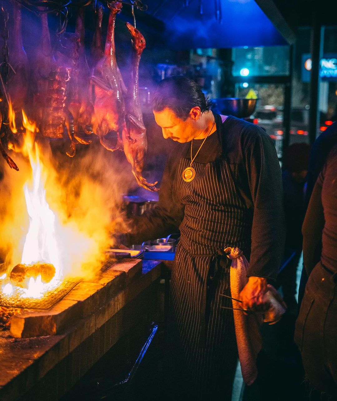 Chef grilling meat over an open fire in a dark setting, showcasing smoke, flames, and traditional cooking techniques.