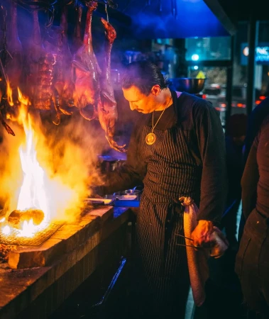Chef grilling meat over an open fire in a dark setting, showcasing smoke, flames, and traditional cooking techniques.