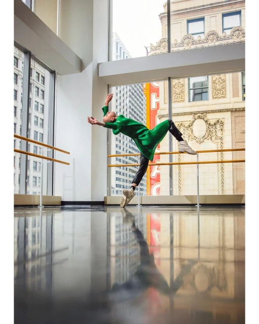 Female ballet dancer in green dress leaps in dance studio with black floor and city view; motion blur and reflection visible.
