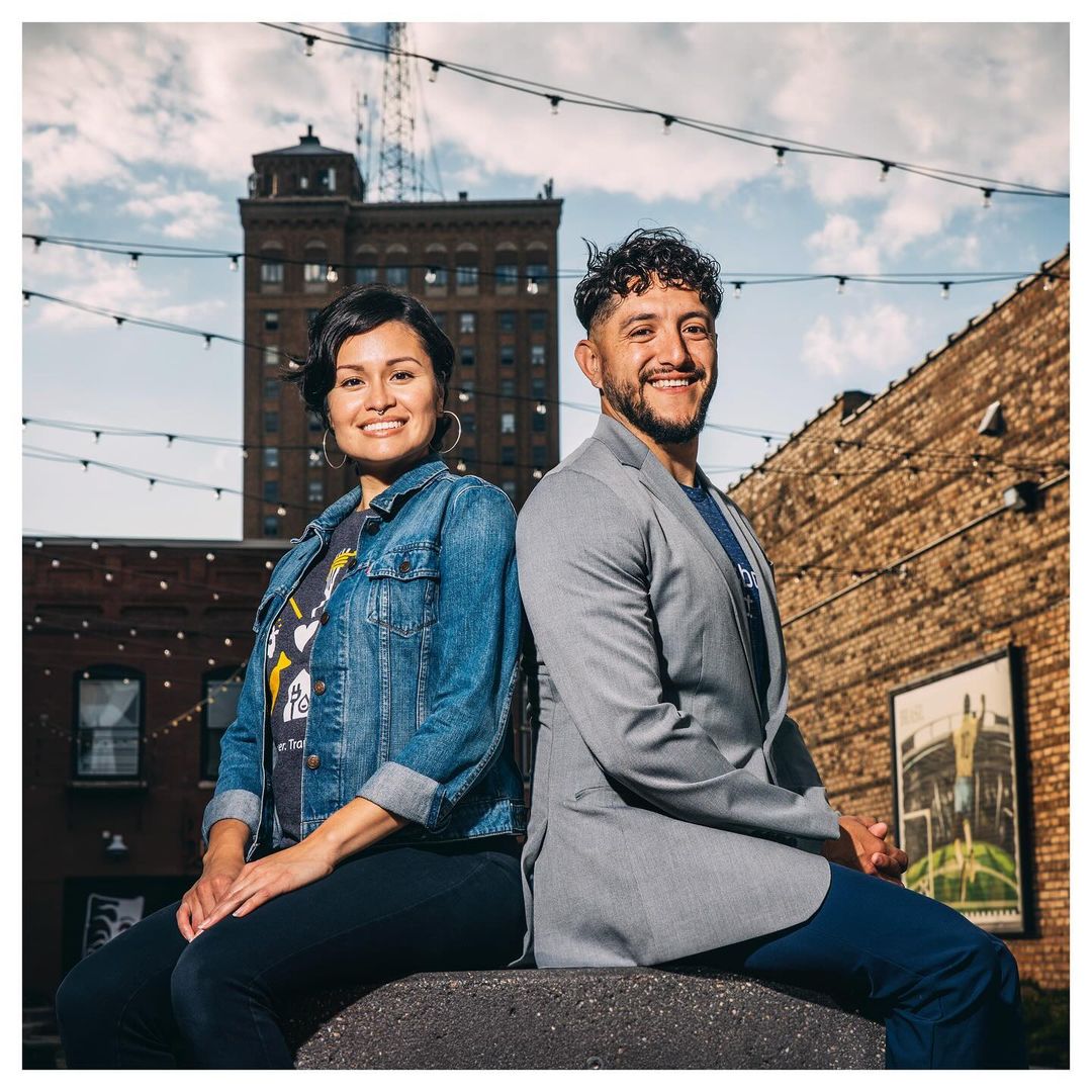 Smiling couple posing outdoors with historic brick buildings in the background on a sunny day with a vibrant blue sky and minimal clouds.