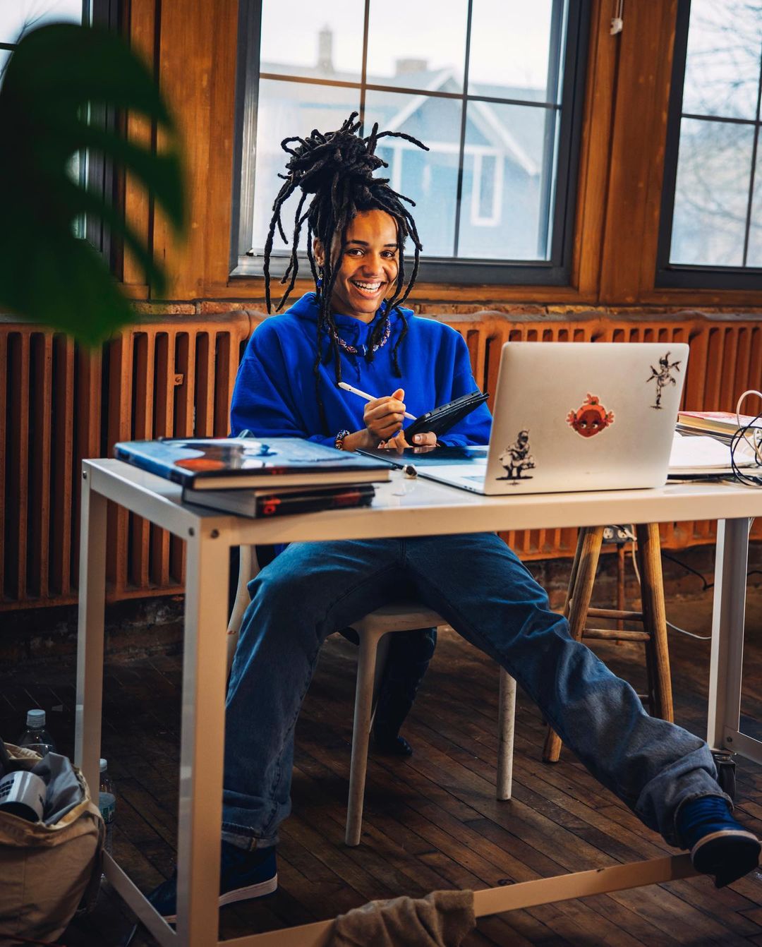 Smiling adult with dreadlocks sitting at a white desk with a laptop, notebook, and pen, working from home in a bright, airy room.