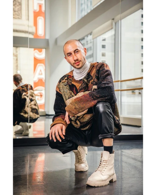 Young adult man squats wearing a printed sweater, black pants, and chunky boots in a dance studio with a reflective wall and windows.
