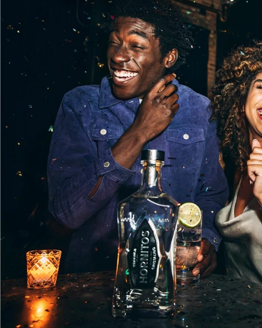 Happy African-American man in blue jacket smiles next to a bottle of Hornitos tequila at a dark bar with a woman and candle.
