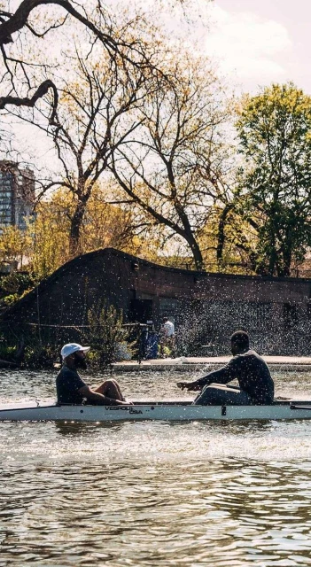 Two people rowing a boat on a river, trees and building in background on a sunny day. Water splashes around the boat as it moves.