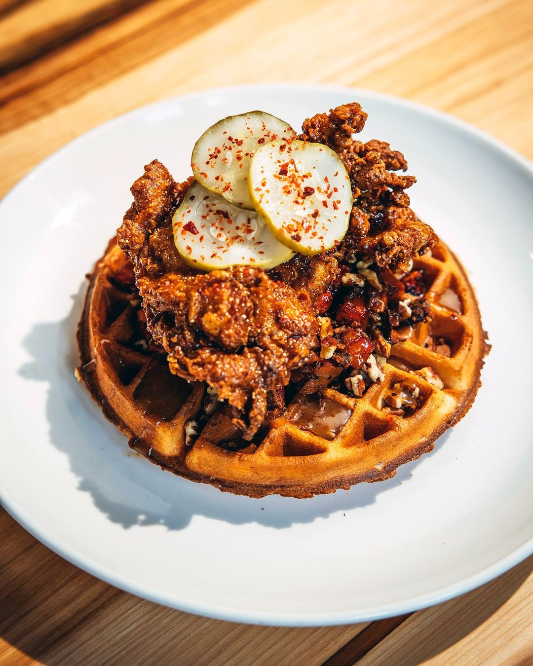 Overhead shot of chicken and waffles on a white plate, garnished with sliced pickles and red pepper flakes, displayed on wood.