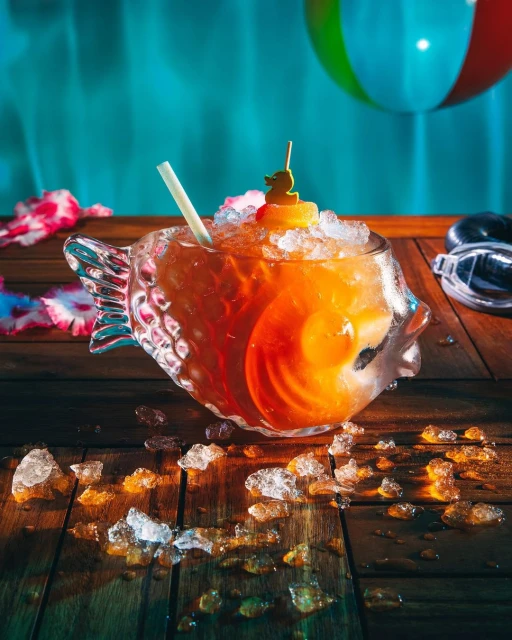 Cocktail in a fish-shaped glass on a wooden table with crushed ice, a straw, and a beach ball in the background. Summer drink.